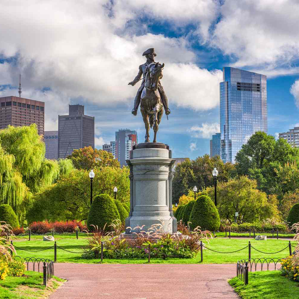 A statue of a man on a horse in a park in Boston, MA with buildings in the distance.