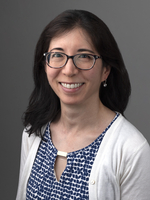 Woman with glasses, white cardigan, and blue patterned shirt smiling.