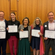 Five people in business attire hold certificates in front of a wood paneled wall.