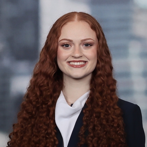 A smiling young woman with curly red hair wearing a white collared shirt and black blazer.