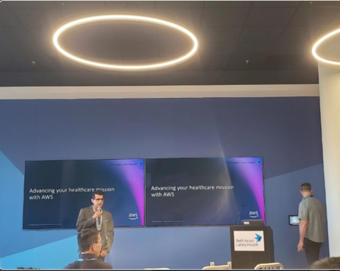 A man speaks at a podium in a conference room with two large monitors behind him.
