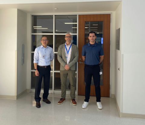 Three men standing in a hallway with white floors and glass doors.