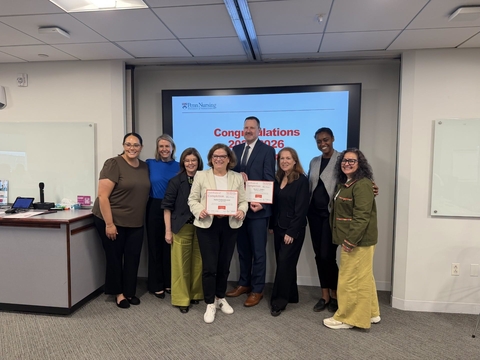 Seven people stand in front of a screen with a congratulations message. A woman holds a certificate.