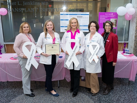 Five women holding pink ribbons and an award plaque stand behind a table.