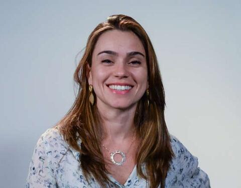 Alice has brown eyes and long, brown hair. She is wearing a blouse. She is standing in front of a white wall and smiling