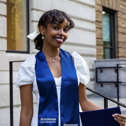 A young woman in a white dress holds a blue diploma while smiling.