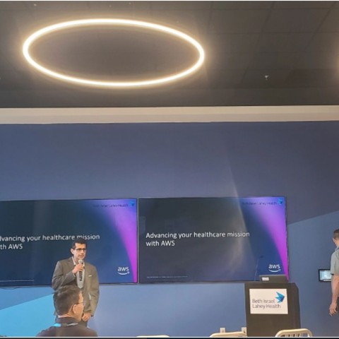 A man speaks at a podium in a conference room with two large monitors behind him.