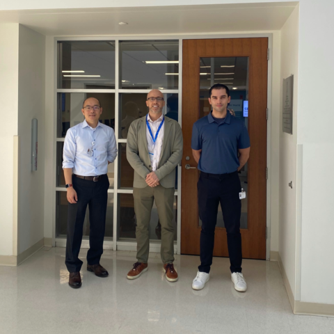 Three men standing in a hallway with white floors and glass doors.