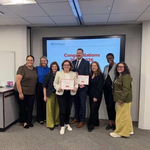 Seven people stand in front of a screen with a congratulations message. A woman holds a certificate.