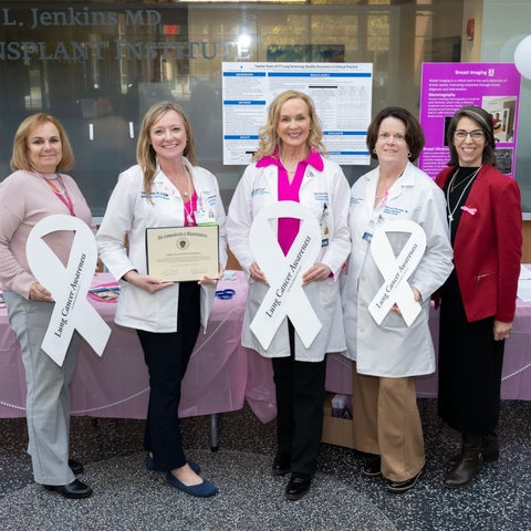 Five women holding pink ribbons and an award plaque stand behind a table.