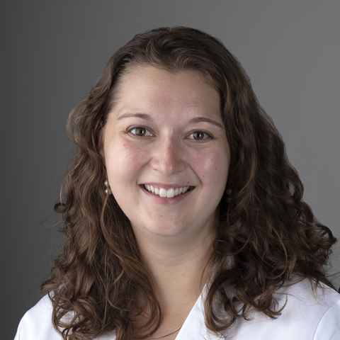 A smiling woman with curly hair wearing a white lab coat.