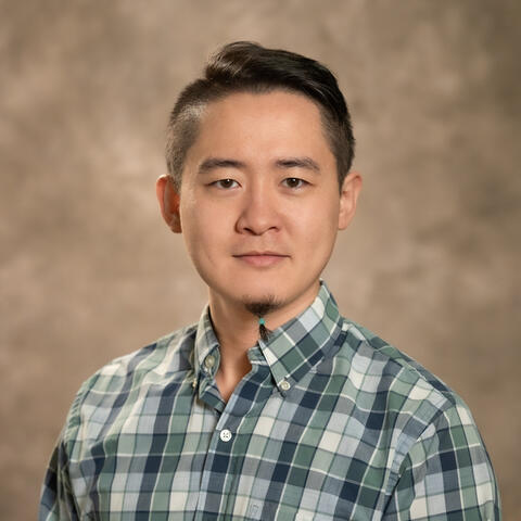 Man in green plaid shirt smiling in front of a brown background