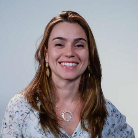 Alice has brown eyes and long, brown hair. She is wearing a blouse. She is standing in front of a white wall and smiling
