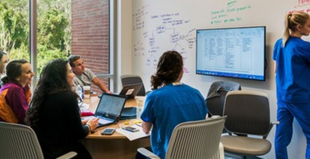 Fellows gathered around a table while another fellow demonstrates/writes on a white board