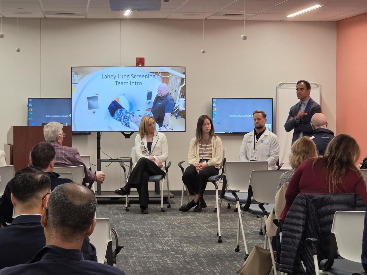 A group of medical professionals discuss in a conference room with a screen showing medical images.