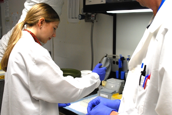 A student in a white coat and blue gloves work at a lab bench.