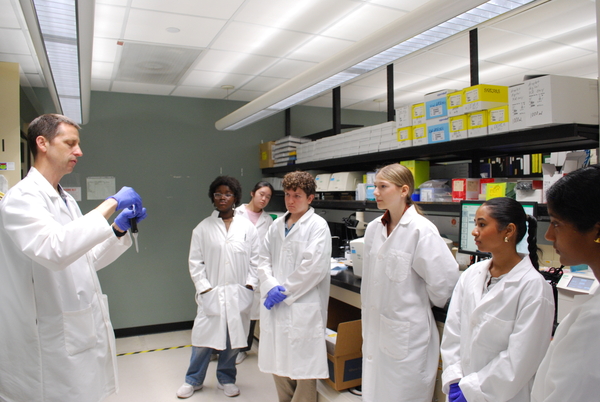 Six students in white coats gather around a lab instructor.