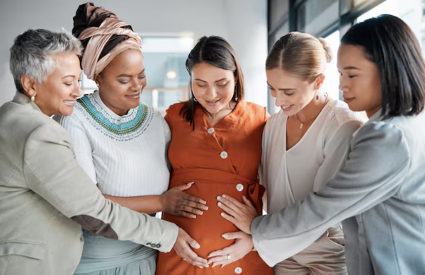 Five women surround a pregnant woman with their hands on her stomach.